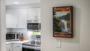 A kitchen with white cabinets, a microwave, a kettle, and a poster of Snoqualmie Falls.