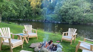 A lakeside setting with a fire pit surrounded by Adirondack chairs, a table with a vase of flowers, and a bottle of hot wood.