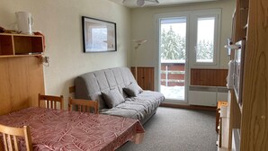 A cozy room with a sofa, a dining table, a framed picture, and a window with a view of trees. - Cozy Studio with Balcony, Near Slopes and Shops, Ski-in/Ski-out Access (Le Pleynet)