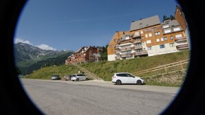 A view through a circular lens of a road with a white car parked on the side. In the background, there are multi-story buildings with balconies, set against a backdrop of mountains and clear sky. - Cozy Studio with Balcony, Near Slopes and Shops, Ski-in/Ski-out Access (Le Pleynet)