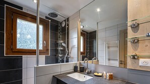 Een moderne badkamer met een grote spiegel, een douche met regendouche, een wastafel met aanrechtblad en een raam met uitzicht op bomen.