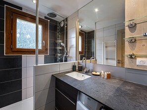 Een moderne badkamer met een grote spiegel, een douche met regendouche, een wastafel met aanrechtblad en een raam met uitzicht op bomen.