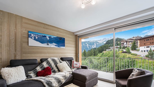 Een moderne woonkamer met een bank, fauteuils en een salontafel. Een groot raam biedt een schilderachtig uitzicht op de bergen.