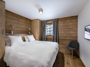 Un dormitorio con pared de madera, una cama, una silla, una ventana con cortinas y una lámpara en el techo.