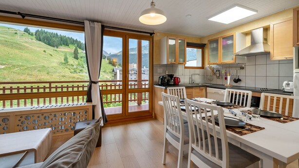 A modern kitchen with wooden cabinets, a dining area with white chairs, and a balcony with a scenic view of a green landscape.
