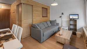A cozy room with a grey sofa, wooden walls, a television, and a dining area with a table and chairs.