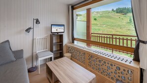 A room with a wooden bench, a TV on a stand, a chair, and a view of a green landscape through a large window.