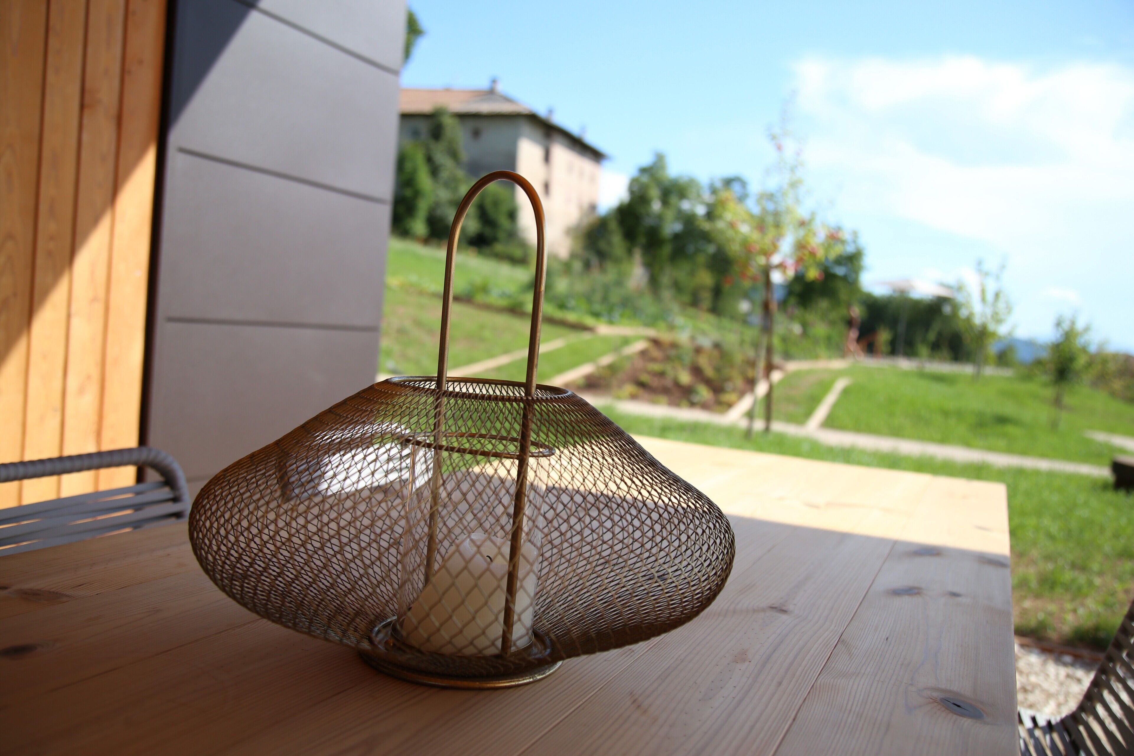 Um candeeiro de vime sobre uma mesa de madeira ao ar livre, com uma casa e vegetação ao fundo.