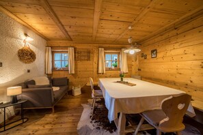 Ein gemütliches Blockhaus-Interieur mit Holzbalkendecke, einem Esstisch mit Stühlen, einer Couch und einer Lampe.