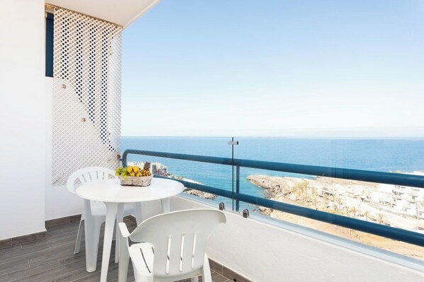 A balcony with a white table and chairs, a basket of fruit, and a view of the sea and buildings. - Home2Book Ocean View Paradise with Pool (Adeje)