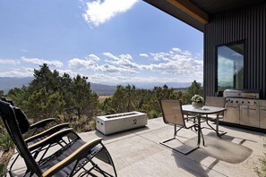 Une terrasse extérieure dotée d’un foyer, de sièges et d’un espace barbecue, le tout offrant une vue sur les montagnes.