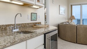 A kitchen with granite countertops, a stainless steel sink, and a built-in oven. There is a beige sofa in the living area with a view of the ocean through large windows.