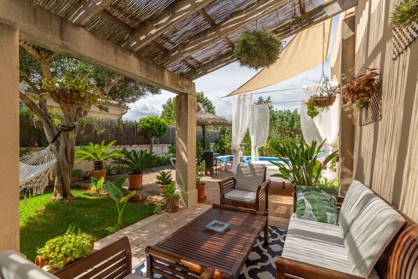 A covered outdoor patio with a wooden table and sofa, surrounded by potted plants and a hammock. - Holiday house with pool very close to the beach, in Playas de Muro, Mallorca (Playa de Muro)