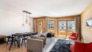 A modern living room with a wooden floor, a dining area with a table and chairs, a sofa, a red chair, and a balcony with a view of buildings. - Charming apartment in Courchevel with parking and Wi-Fi included (Courchevel 1650)