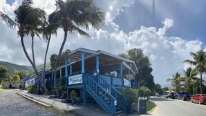 A small, two-story building with a blue exterior and a covered porch, located at the end of a road with parked cars and palm trees in the background. - Walk to Cane Bay Beach Cottage - End Unit with Mountain Views (Christiansted)