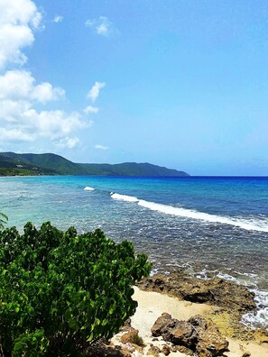 A beach with clear blue water, rocky shoreline, and green vegetation. - Walk to Cane Bay Beach Cottage - End Unit with Mountain Views (Christiansted)
