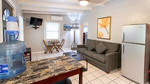 A compact living space with a kitchenette, a large water dispenser, a sofa, a TV mounted on the wall, and a framed picture. - Walk to Cane Bay Beach Cottage - End Unit with Mountain Views (Christiansted)