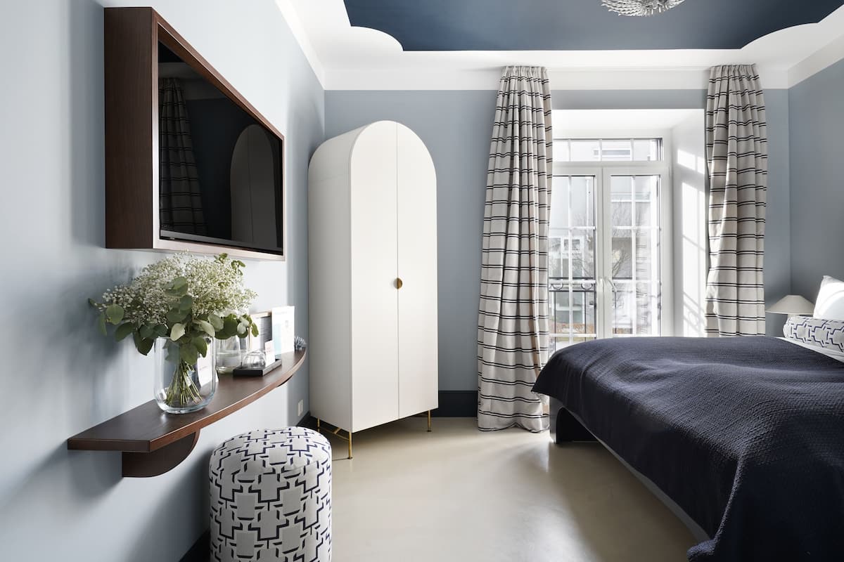 Une chambre à coucher comprenant un lit, une armoire, un miroir, une étagère avec un vase et une fenêtre avec des rideaux.