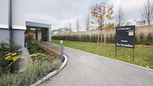 Une entrée moderne d’un bâtiment, avec une enseigne indiquant l’hôtel « The Maximilian », un chemin et des espaces verts aménagés.