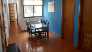 A room with a glass-top dining table, wooden chairs, a window with blinds, and a blue wall. - Ipanema Beach (Rio de Janeiro)
