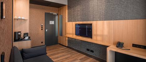 A modern hotel room with a wooden floor, a sofa, a desk with a phone, and a wall-mounted screen displaying a list.