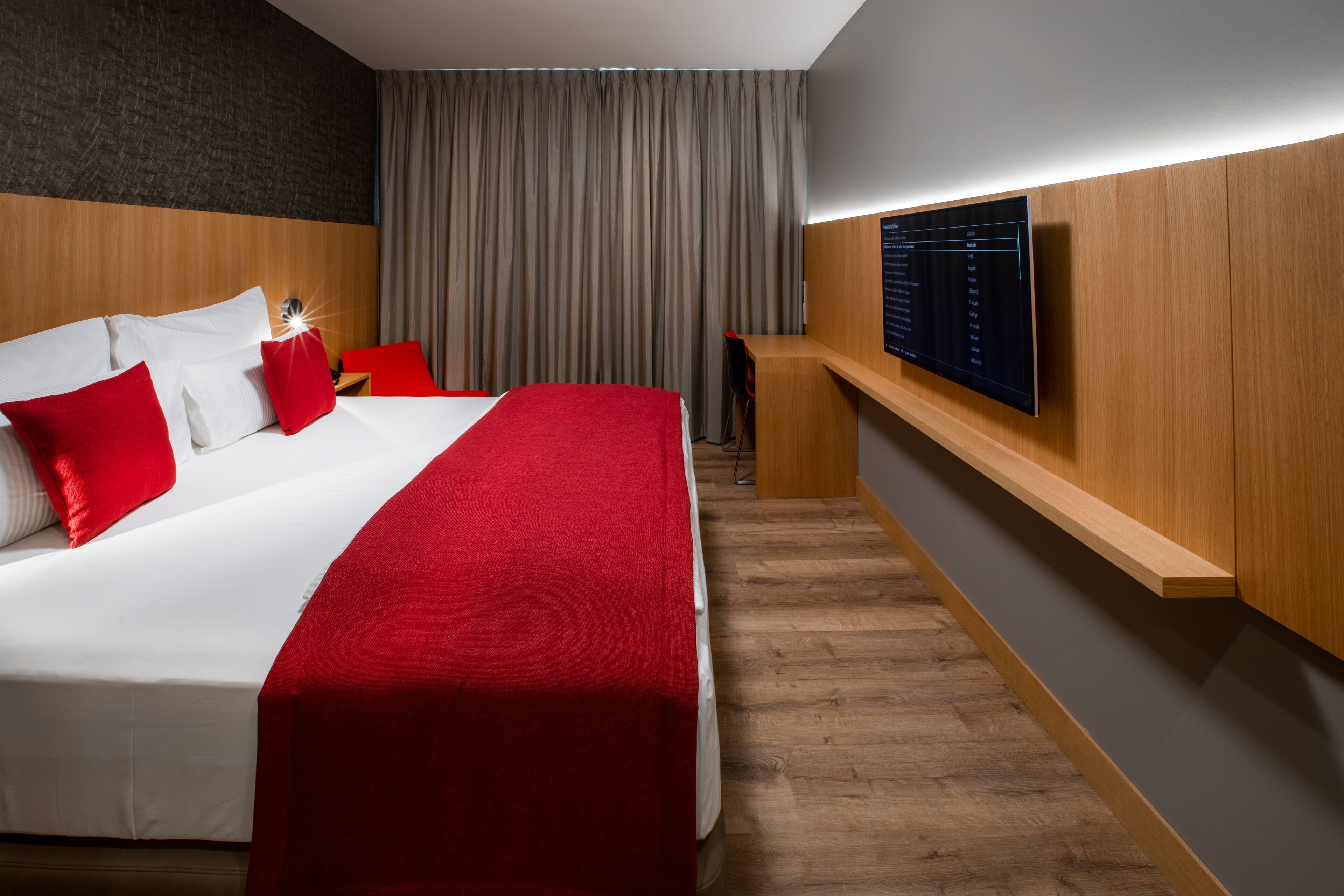 A modern hotel room with a large bed, red and white bedding, a wooden headboard, a flat-screen TV mounted on the wall, and a wooden desk with a chair.