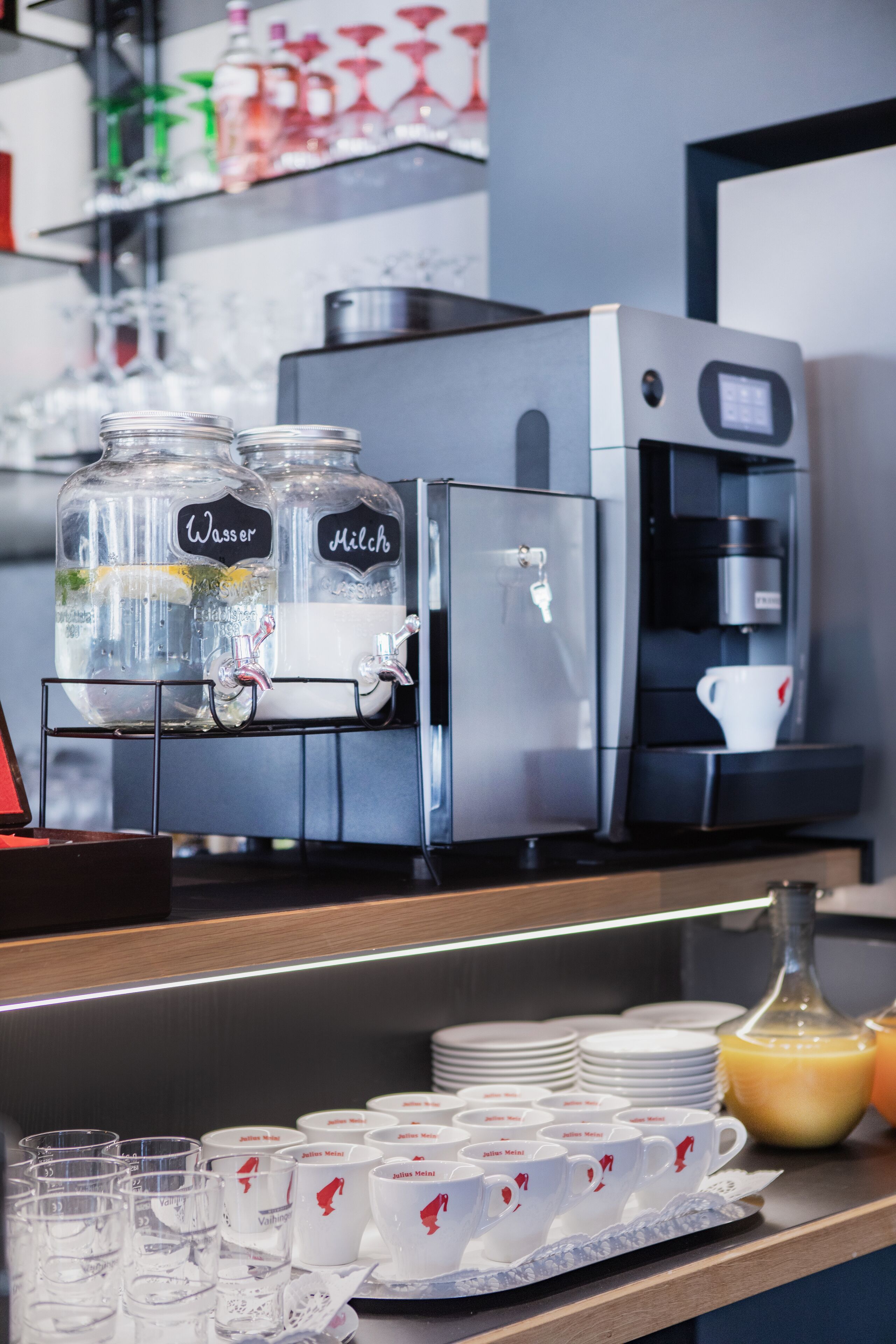 A coffee station with a coffee machine and two water dispensers, a tray of cups, and a bottle of orange juice.
