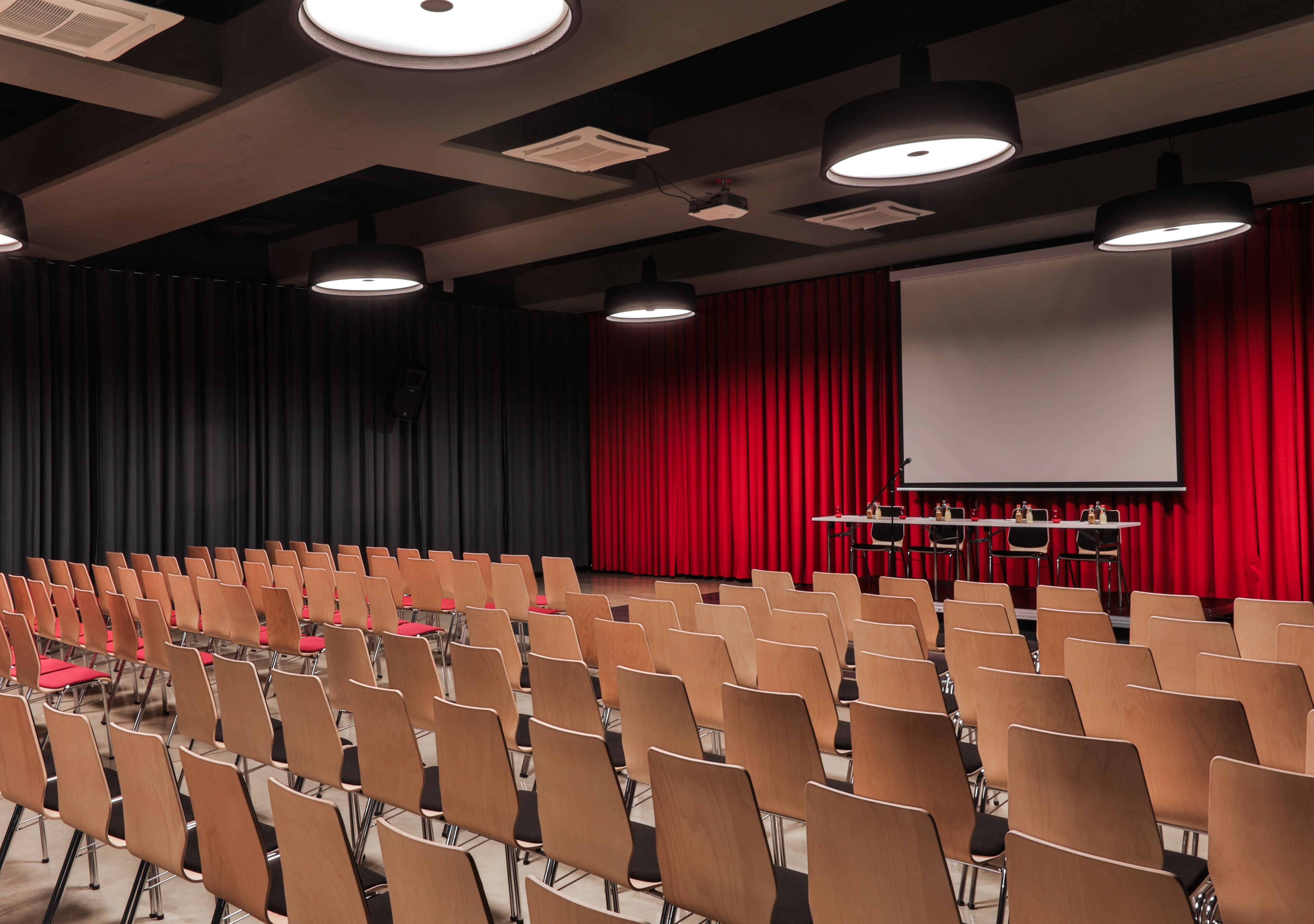 A conference room with rows of chairs, a stage with a projector screen, and red curtains.
