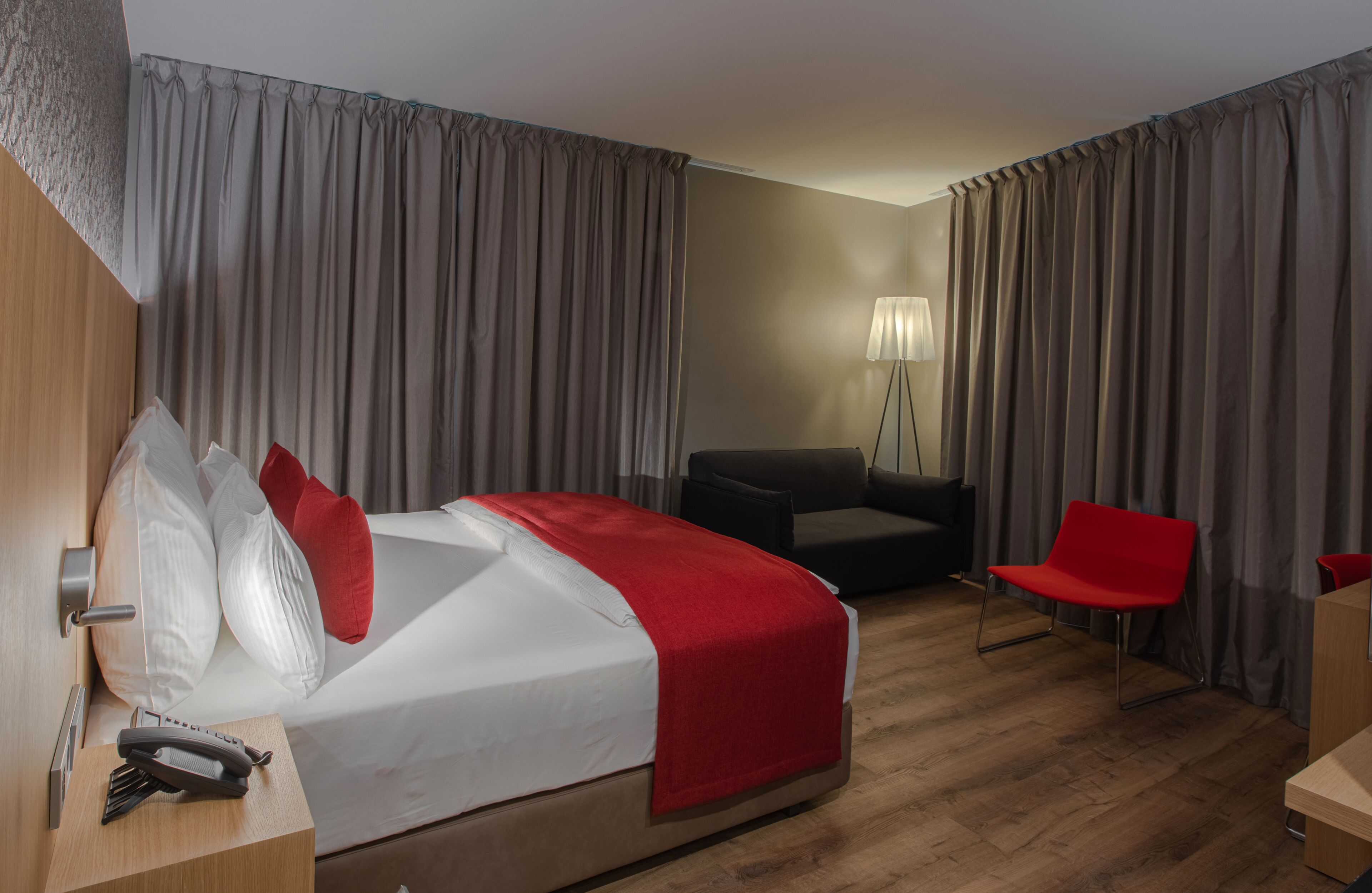 Ett hotellrum med en säng, ett skrivbord med en telefon, en röd stol, en soffa och en lampa.