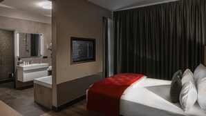 A modern hotel room with a large bed, a bedside table, a red throw blanket, a wall-mounted TV, and a bathroom with a mirror, sink, and towel rack. - Ocak Hotel (Berlin)