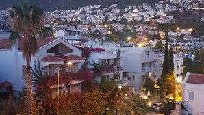 Un pueblo costero con vistas al mar y a las montañas. Se observan edificios blancos con balcones, algunos adornados con flores. Se distingue una zona pavimentada con sillones de playa y sombrillas.