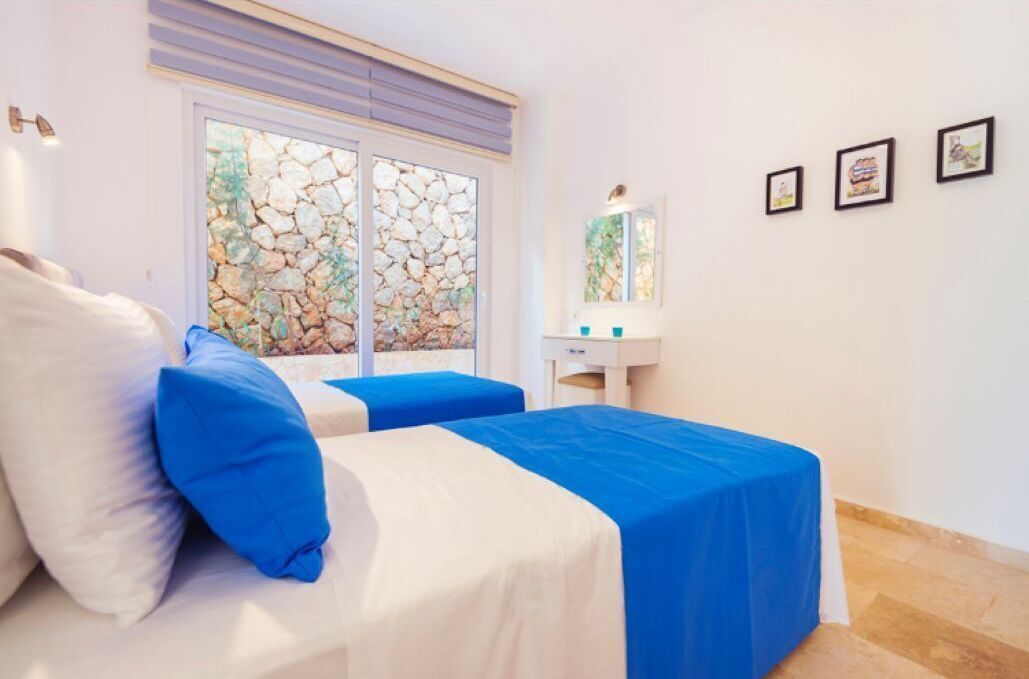 A bedroom with two beds, a stone wall, a window, a mirror, and three framed pictures on the wall.