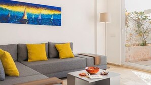 Un salón moderno con un sofá gris, cojines amarillos, un cuadro de veleros, una lámpara de pie y vistas al exterior.
