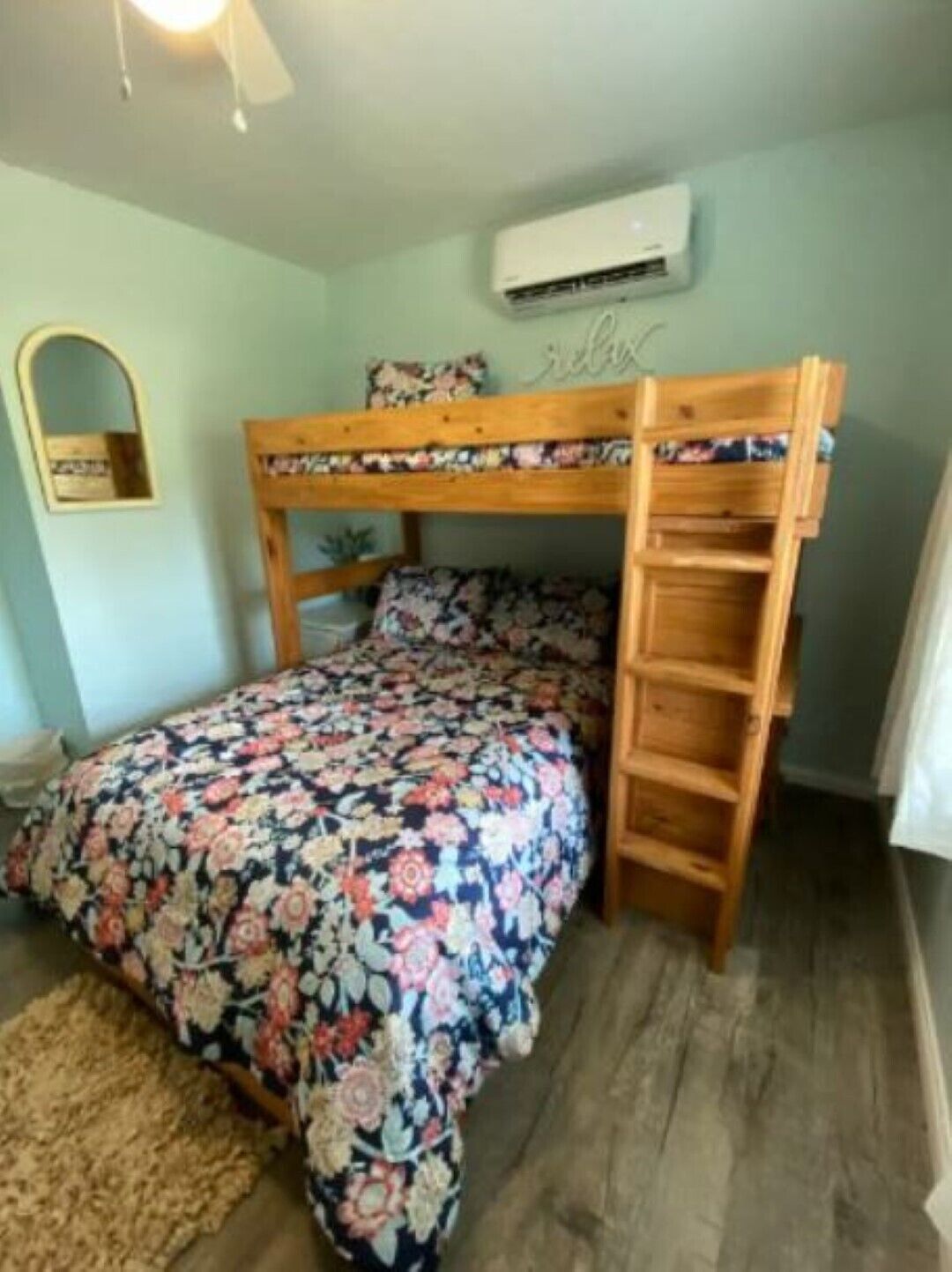 Un lit superposé avec une couvre-lit à motifs floraux, un petit miroir et un appareil de climatisation fixé au mur.