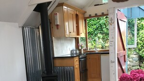 A kitchen with wooden cabinets, a black stove, and a wooden dining table with a chair. - Fisherman's Studio in Cadgwith Cove - sleeps 2 guests  in 1 bedroom (HELSTON)