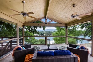 A covered outdoor seating area with wicker furniture and ceiling fans, overlooking a lake and forested landscape. - The Gathering Place on Lake LBJ (Burnet)