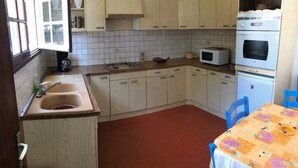 Eine kleine Küche mit Spüle, Herd, Mikrowelle und Backofen. Es gibt ein Fenster mit Jalousien, einen Tisch mit einer blumigen Tischdecke und blaue Stühle.