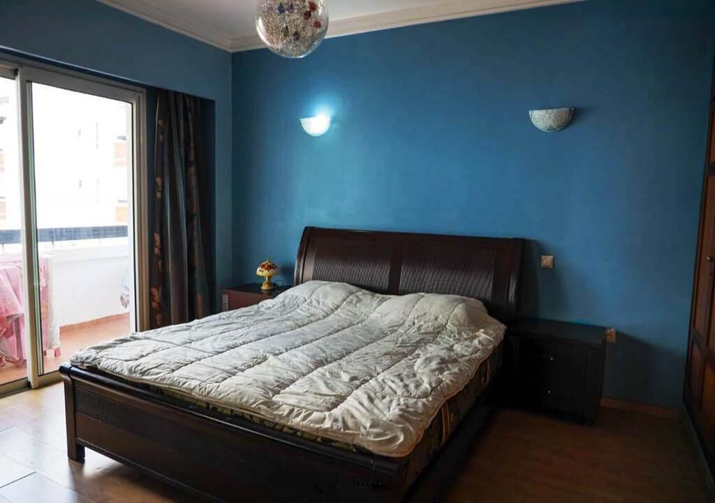 A bedroom with a bed, a nightstand, a wall-mounted lamp, and a view through a sliding door to an outdoor balcony.