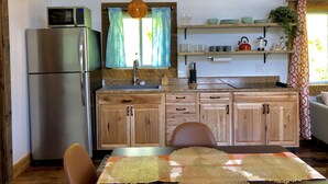 Una cocina con armarios de madera, un refrigerador de acero inoxidable, un microondas y un lavamanos. Hay dos ventanas con cortinas, una lámpara colgante y una repisa con vajilla y un hervidor.