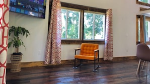 Una habitación con un televisor grande montado en la pared mostrando un canal deportivo, dos ventanas con cortinas, una planta en maceta y suelo de madera.