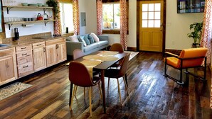 Un amplio espacio habitable con una mesa de comedor, sillas y un sofá. Hay una cocina con armarios de madera y una encimera. El cuarto tiene piso de madera y ventanas grandes con cortinas estampadas.