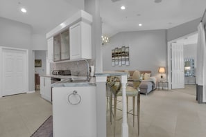A modern kitchen with a central island, a dining area with a sofa and chairs, and a living room with a couch and a lamp. - HAWK'S NEST 202 - Luxurious Remodeled Fiddler's Creek Coach Home (Naples)