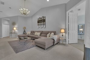 A modern living room with a sectional sofa, a glass coffee table, a chandelier, and a wall art piece.