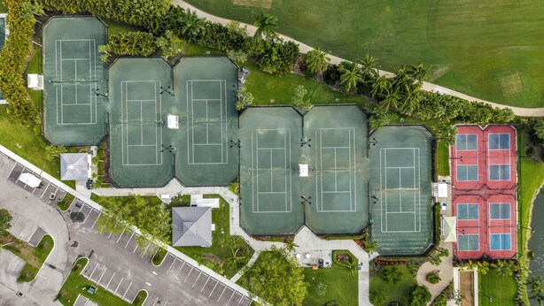 Vista aérea de un complejo deportivo con varias canchas de tenis, zonas de estacionamiento y áreas verdes circundantes.