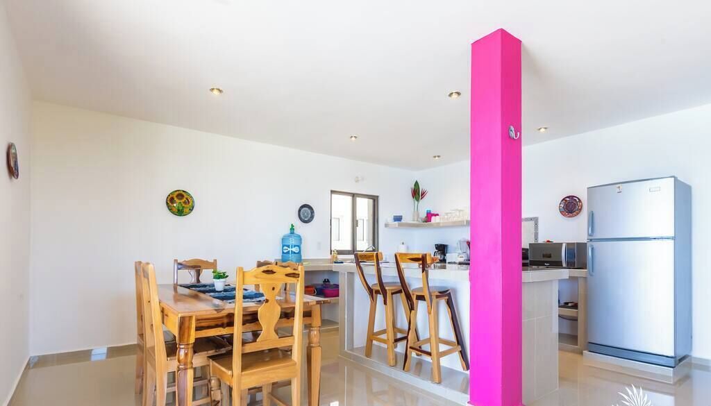 A modern kitchen with wooden chairs, a dining table, and a bright pink accent wall.