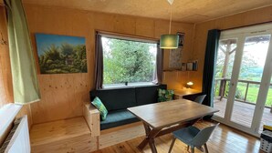 Ein gemütliches Zimmer mit Holzboden, einem Esstisch mit Stühlen, einer Bank mit Kissen und Vorhängen am Fenster. Blick ins Grüne.