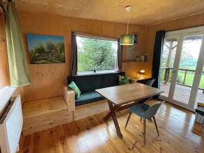 A cozy room with a wooden floor, a dining table with chairs, a bench with cushions, a window with curtains, and a view of greenery outside.