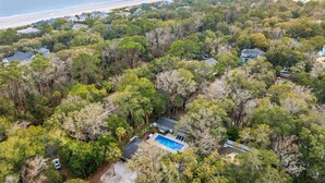 An aerial view of a residential area with a swimming pool, surrounded by dense trees and a beach in the background. - 12 Park Rd - Octopus Oasis (Hilton Head Island)