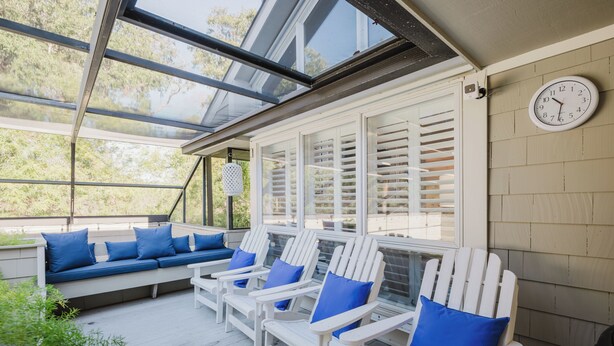 A covered outdoor seating area with white Adirondack chairs and blue cushions, a white clock on the wall, and a view of trees through the glass ceiling. - 4 Firethorn Way - 6 Bedroom, 3rd Row, South Forest Beach Home (Hilton Head Island)