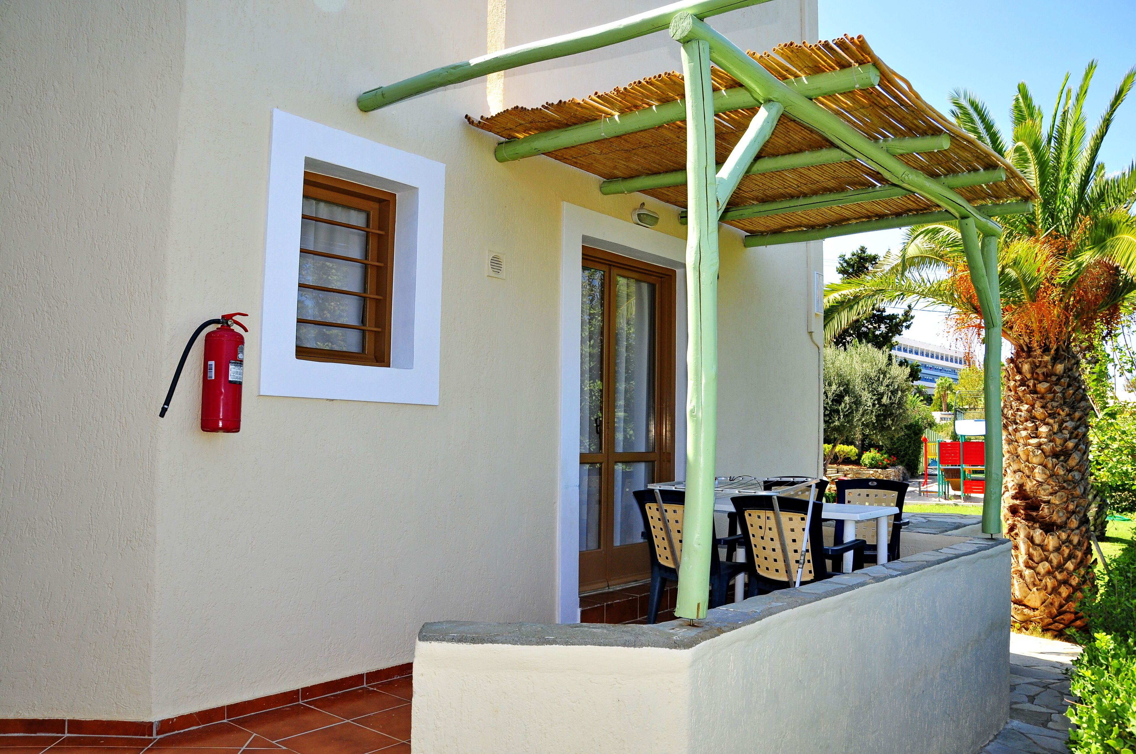A covered outdoor seating area with tables and chairs, a fire extinguisher mounted on the wall, and a palm tree in the background.